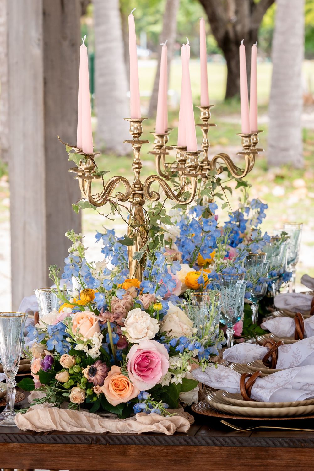 Elegant table setting with brass candelabra, pink and blue flowers, and place settings on cream linens.