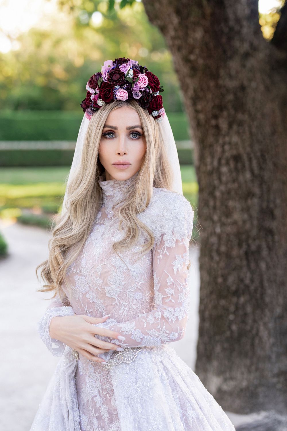 A figure in a white lace dress and purple flower crown poses outdoors in a dreamy garden setting.