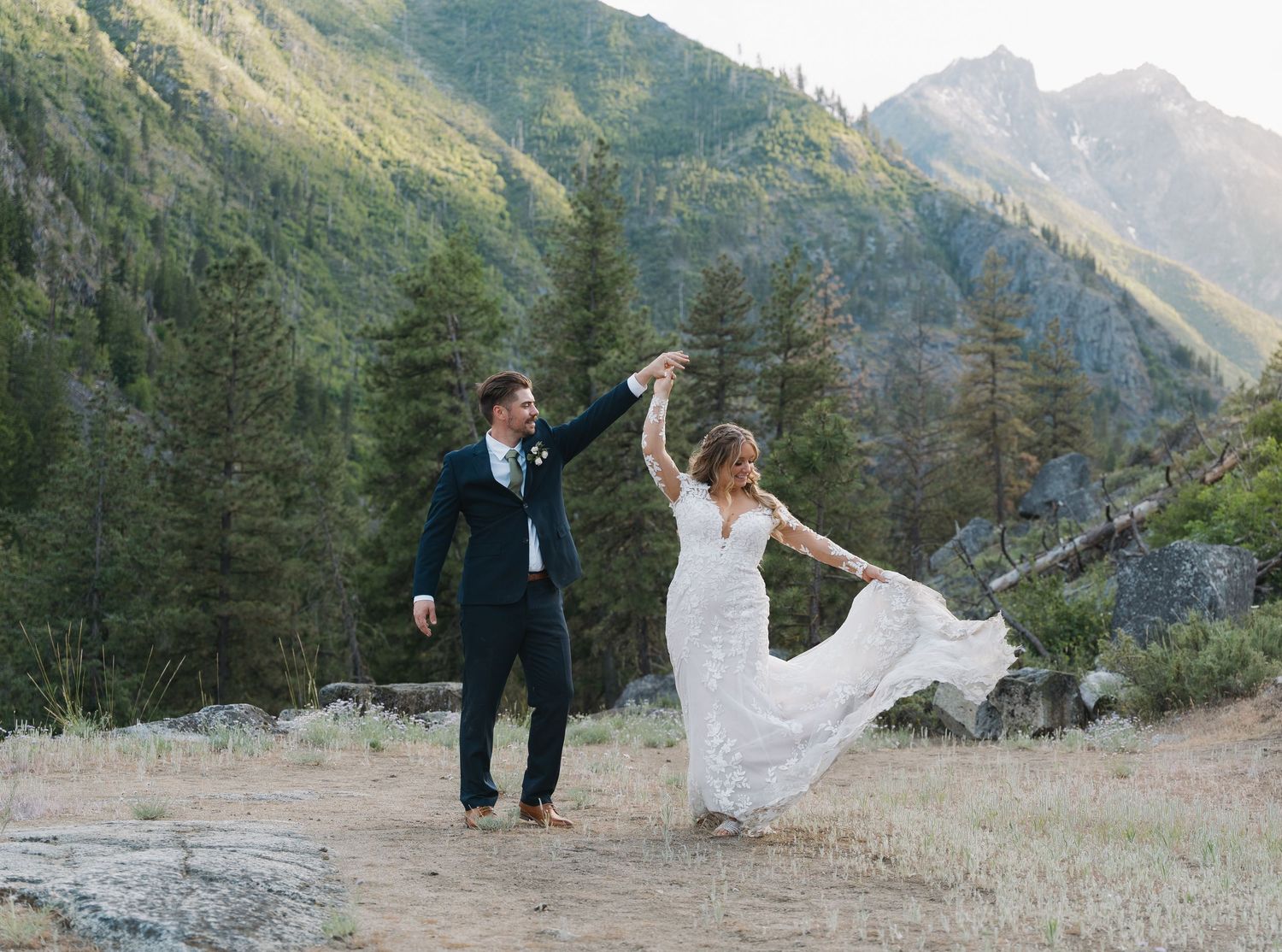 A couple dances joyfully on a mountain trail surrounded by evergreen trees and dramatic peaks in the background.