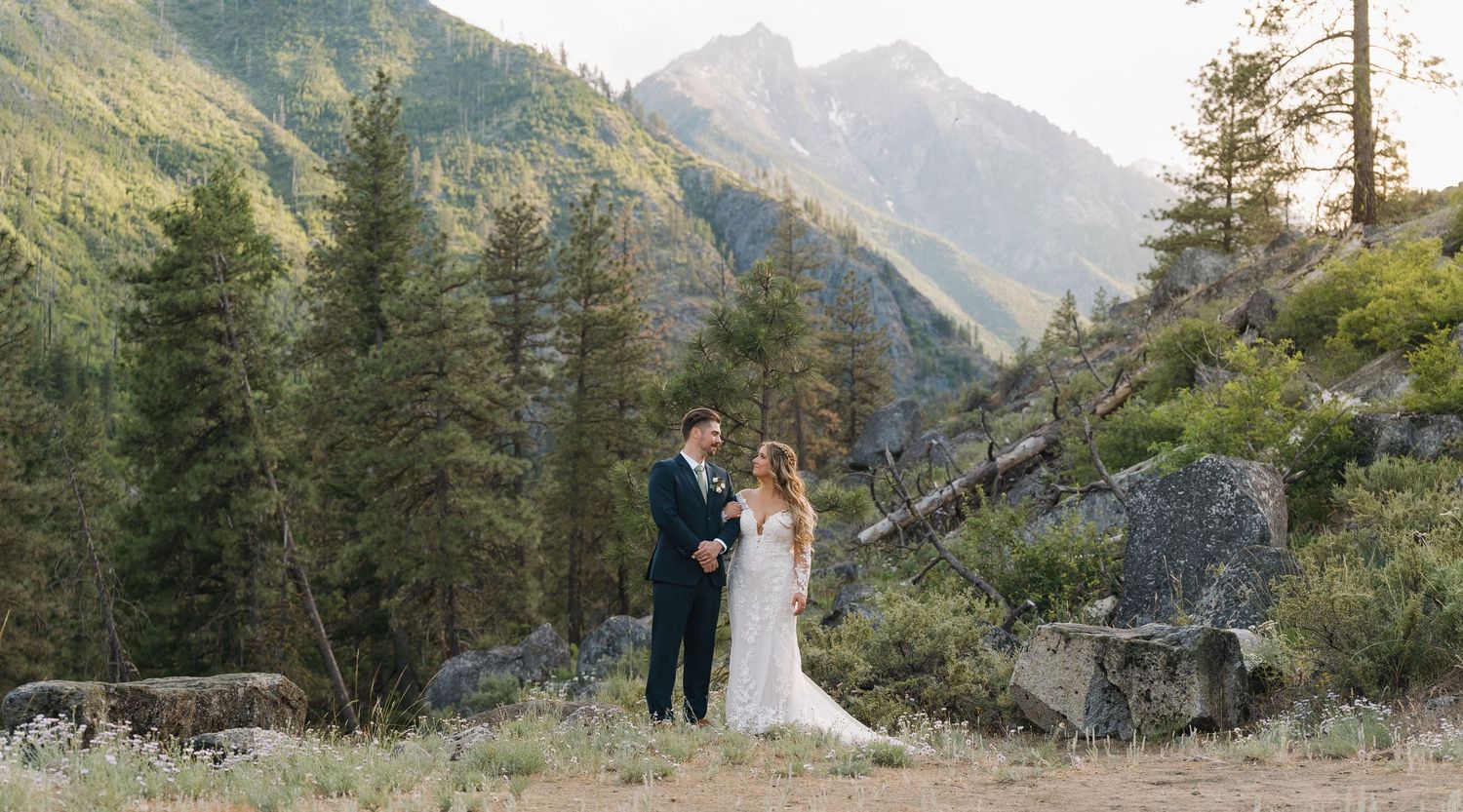 A couple in wedding attire stands together in a scenic mountain landscape with evergreen trees and rocky terrain.