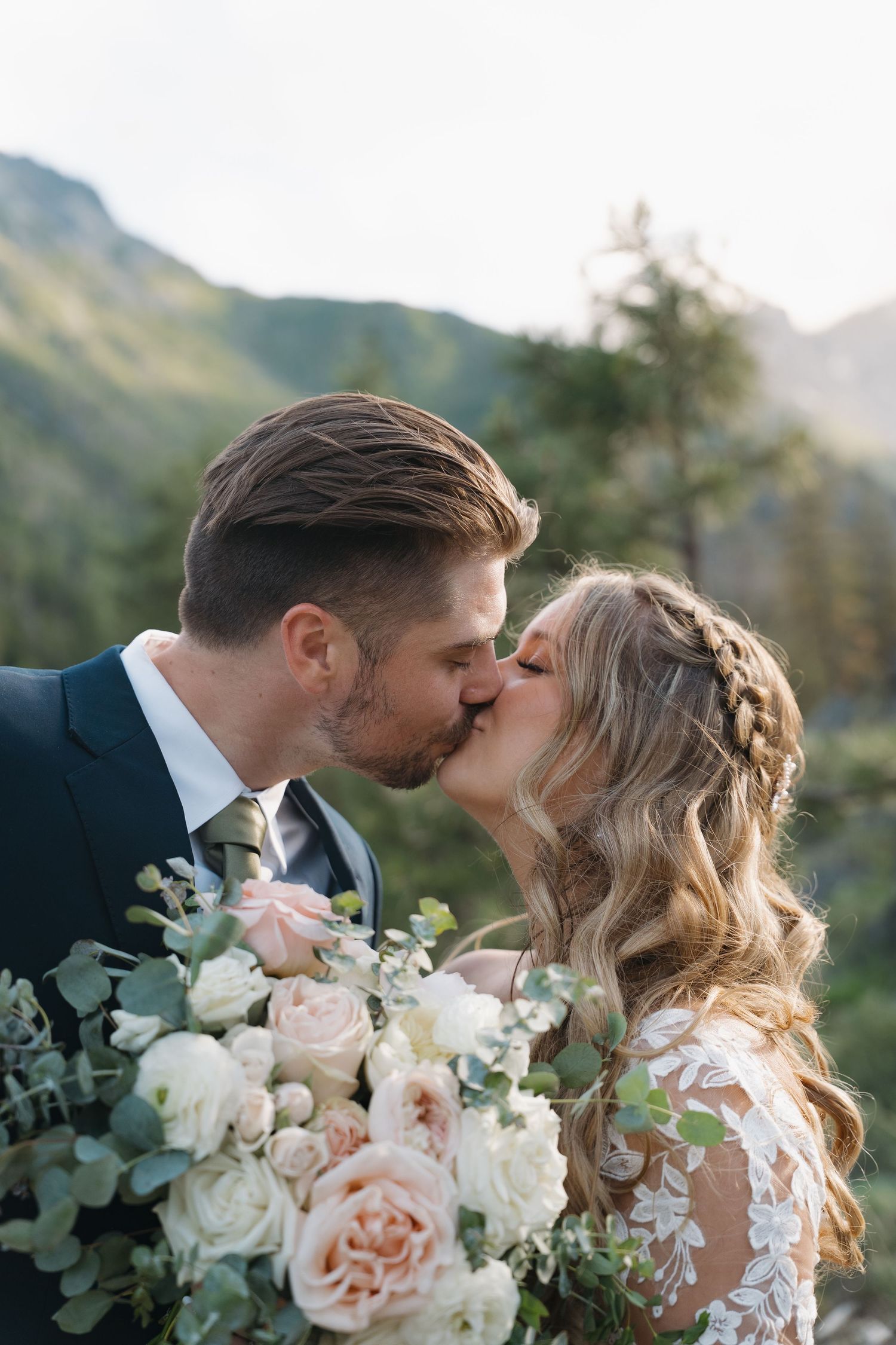 A romantic wedding couple shares an intimate kiss outdoors with mountains and greenery in the background.