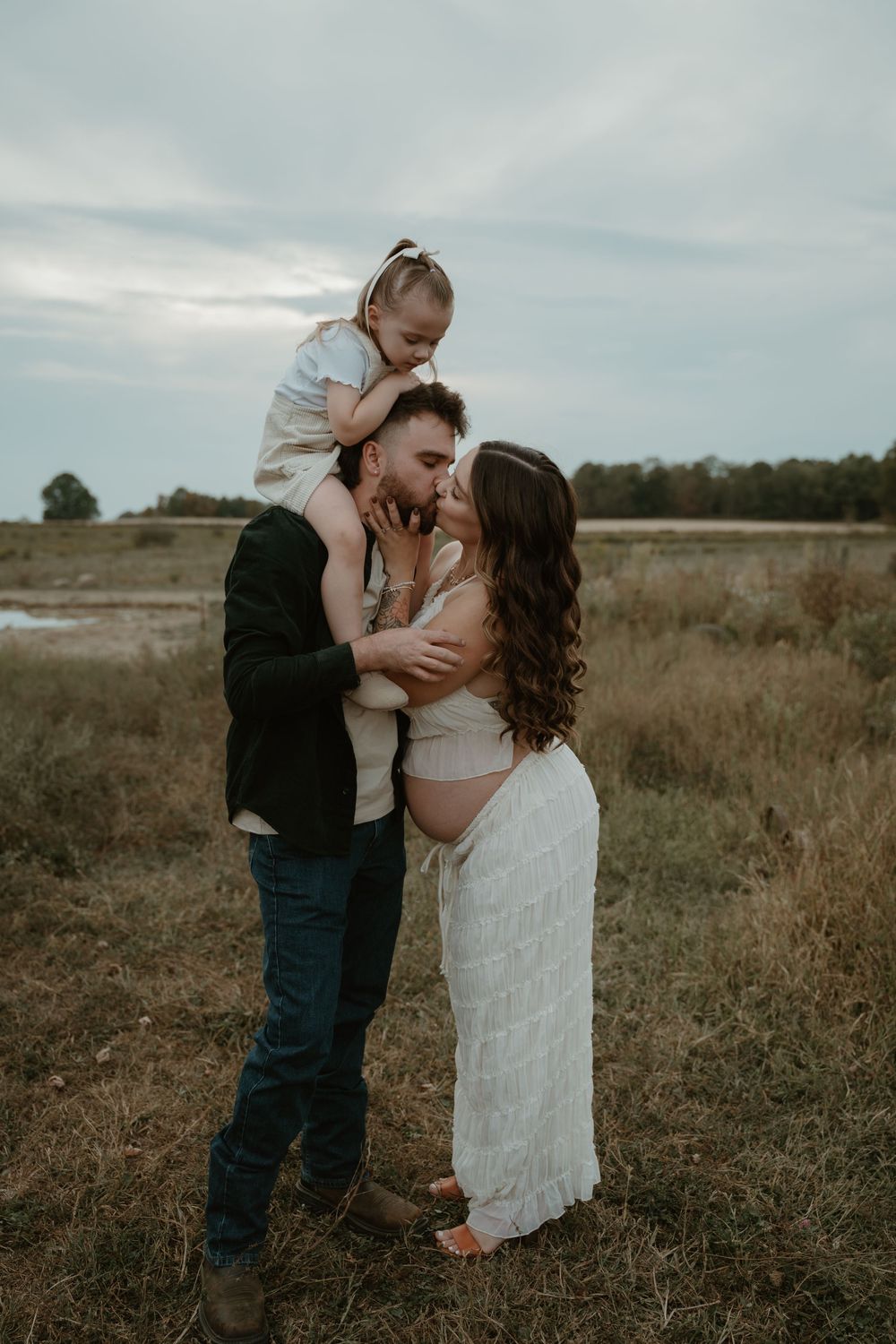 Expecting family of three sharing a kiss outdoors in a field, with toddler daughter sitting on father's shoulders.