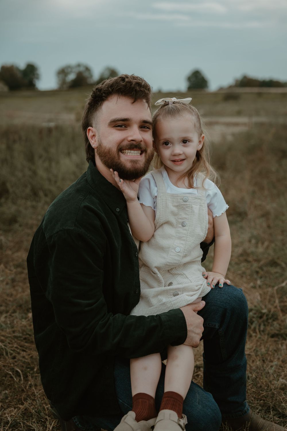 Smiling bearded father sitting in autumn field holding his young toddler daughter in white overalls and bow headband.