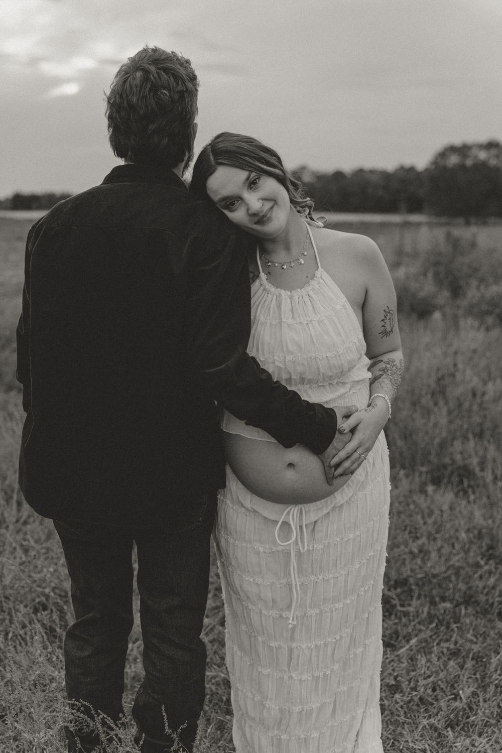 Black and white photo of pregnant woman in white dress leaning on partner in field.