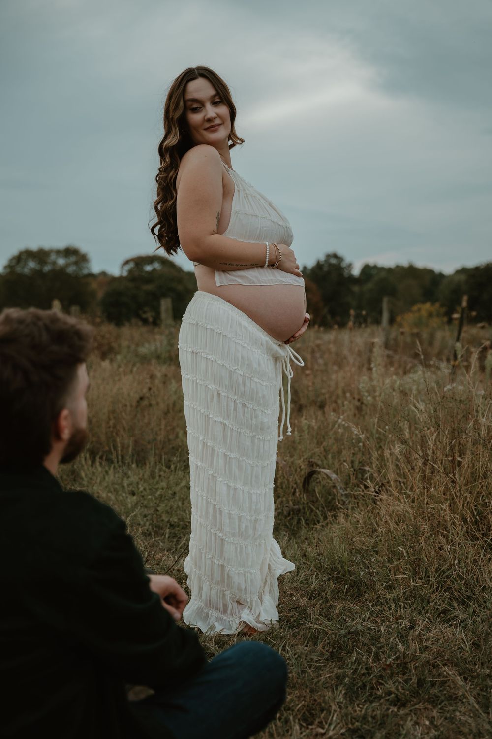Pregnant woman in white fringe dress smiling in autumn field with partner seated nearby.