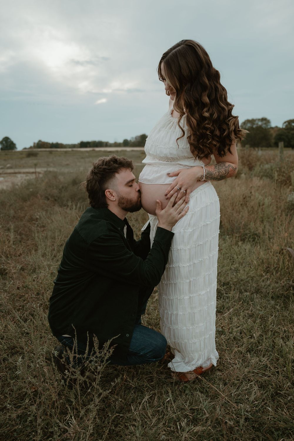 Man kneeling to kiss pregnant woman's baby bump in golden field at dusk.