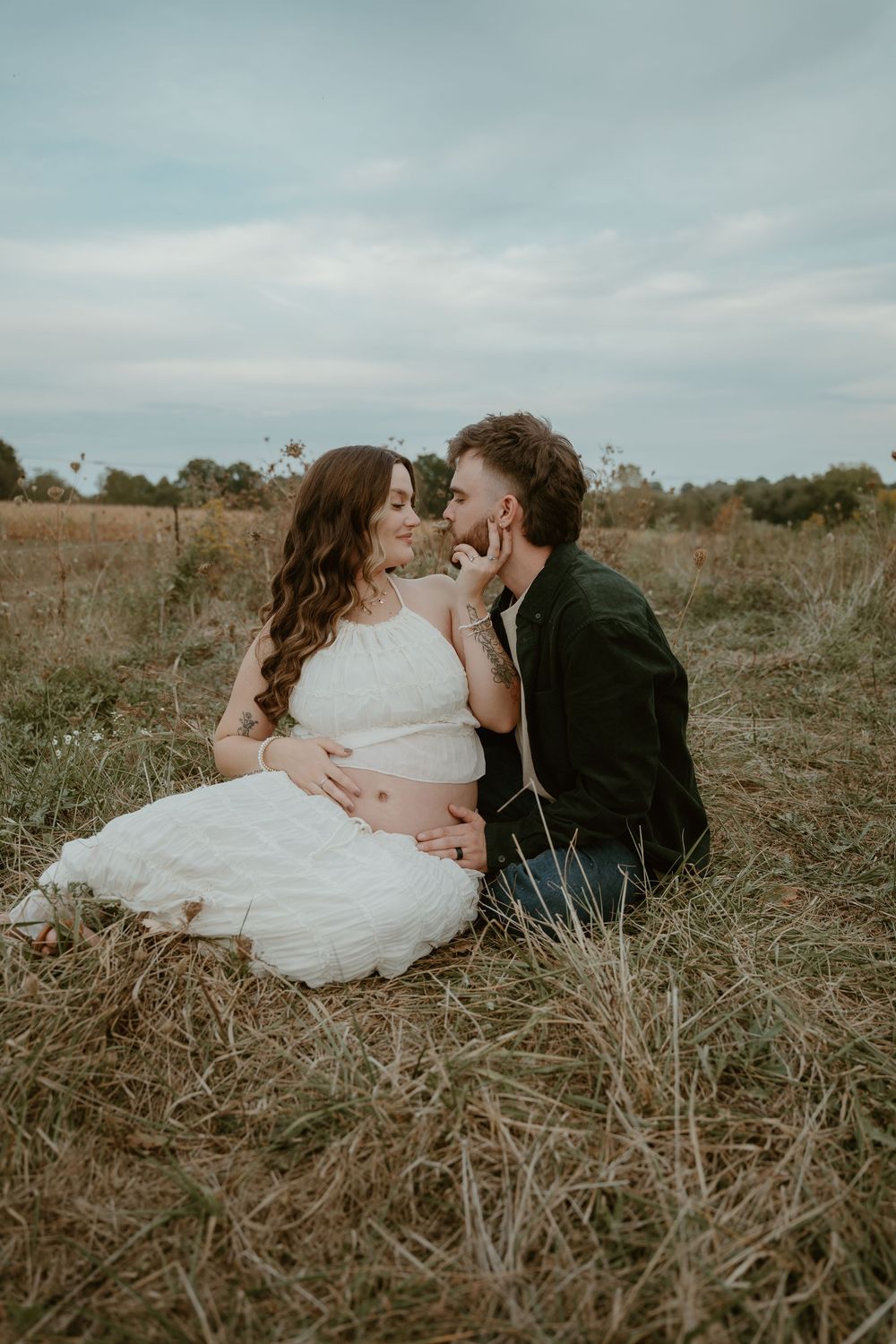 Expecting couple sitting in dry grass field gazing lovingly at each other.