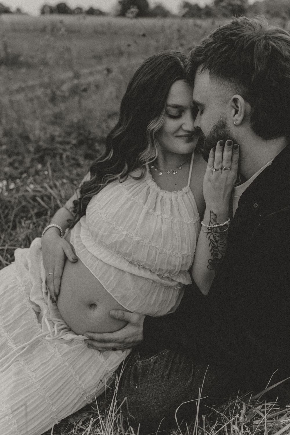 Black and white maternity photo of couple embracing in field, hands on baby bump.