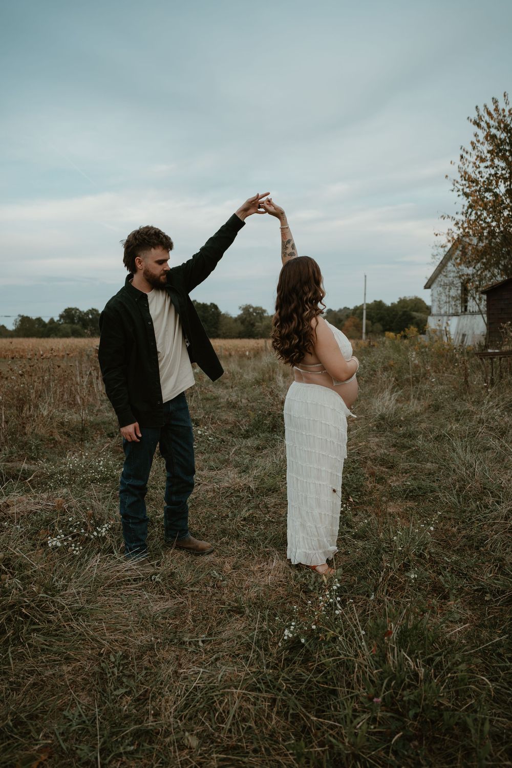 Man spinning pregnant partner by hand in rustic autumn field near old barn.