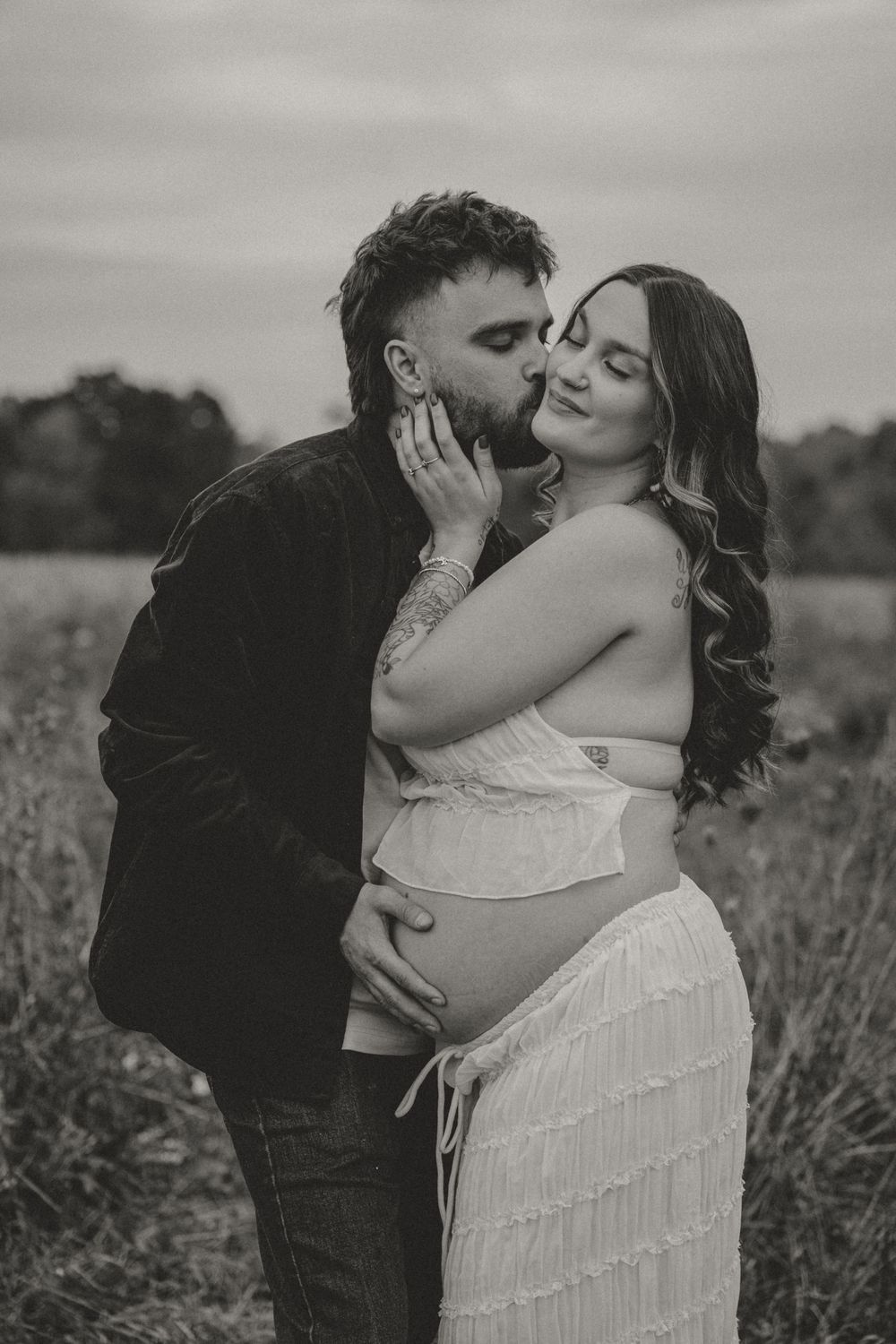 Black and white photo of man kissing pregnant woman's cheek in open field.