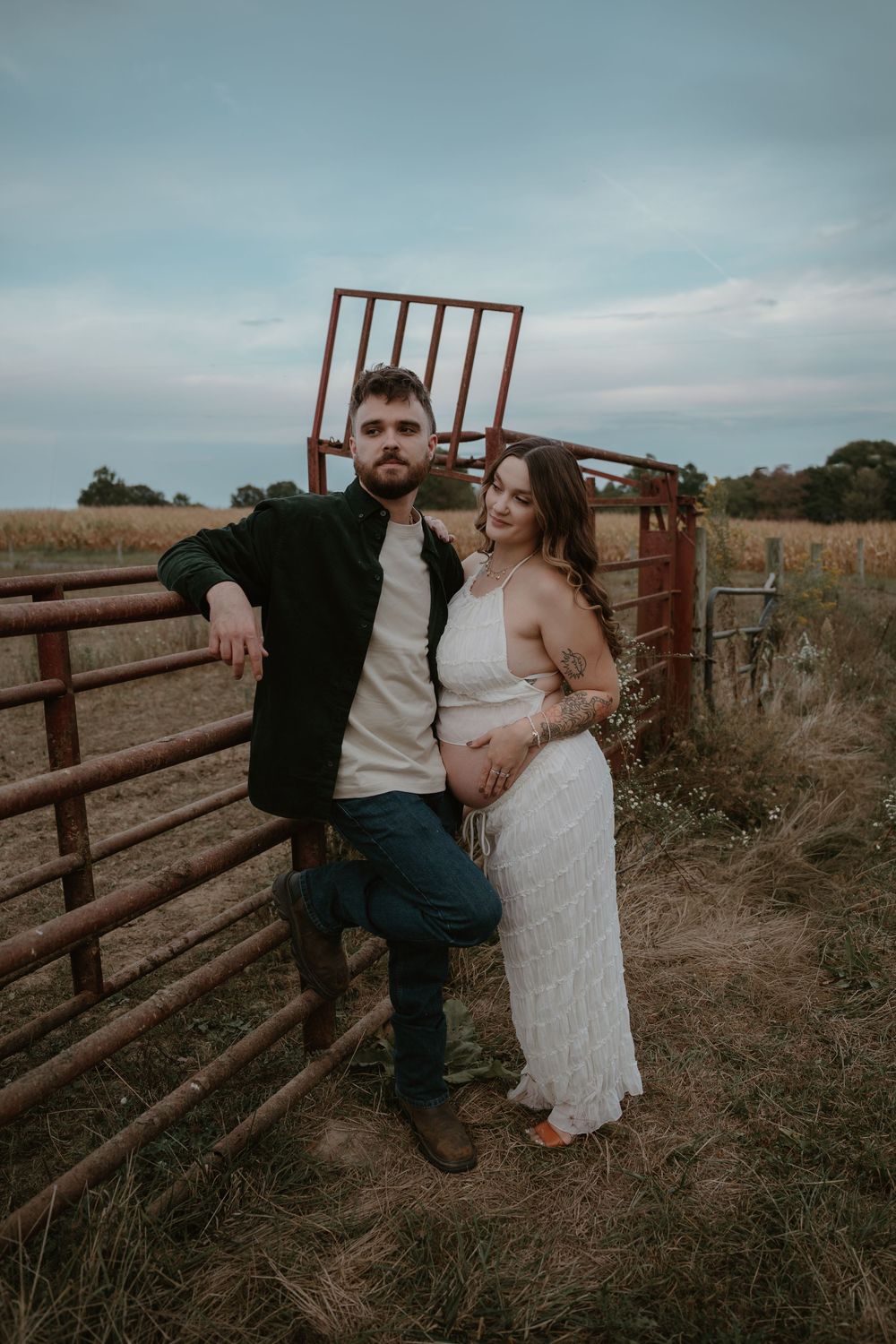 Expecting couple posing by rustic red farm gate in open countryside field.