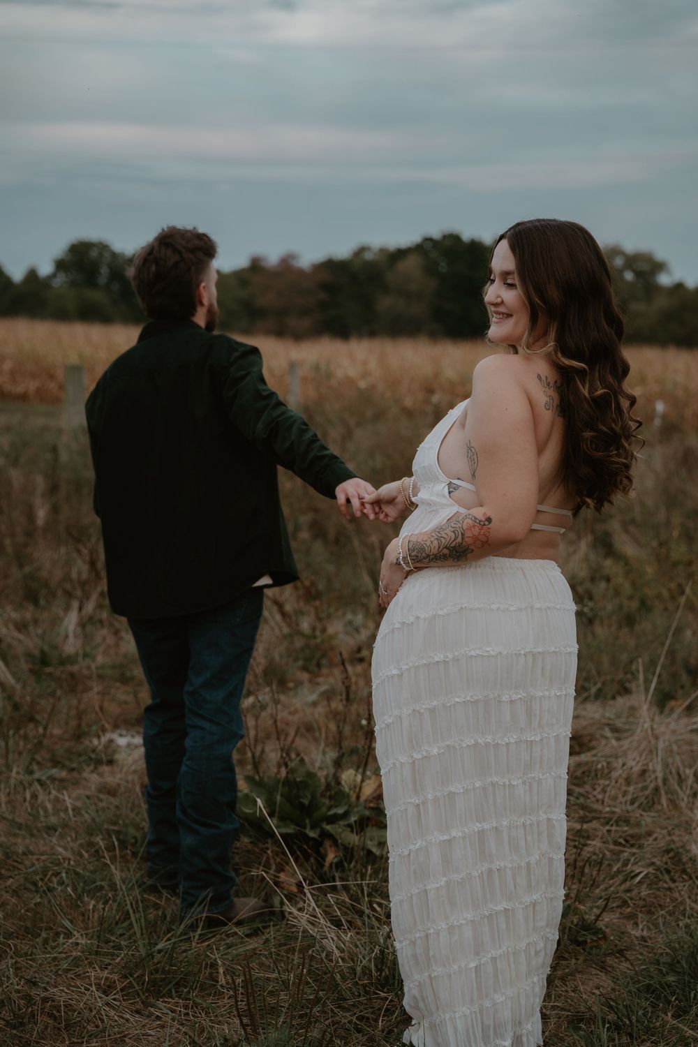 Smiling pregnant woman in white dress holding hands with partner in autumn field.