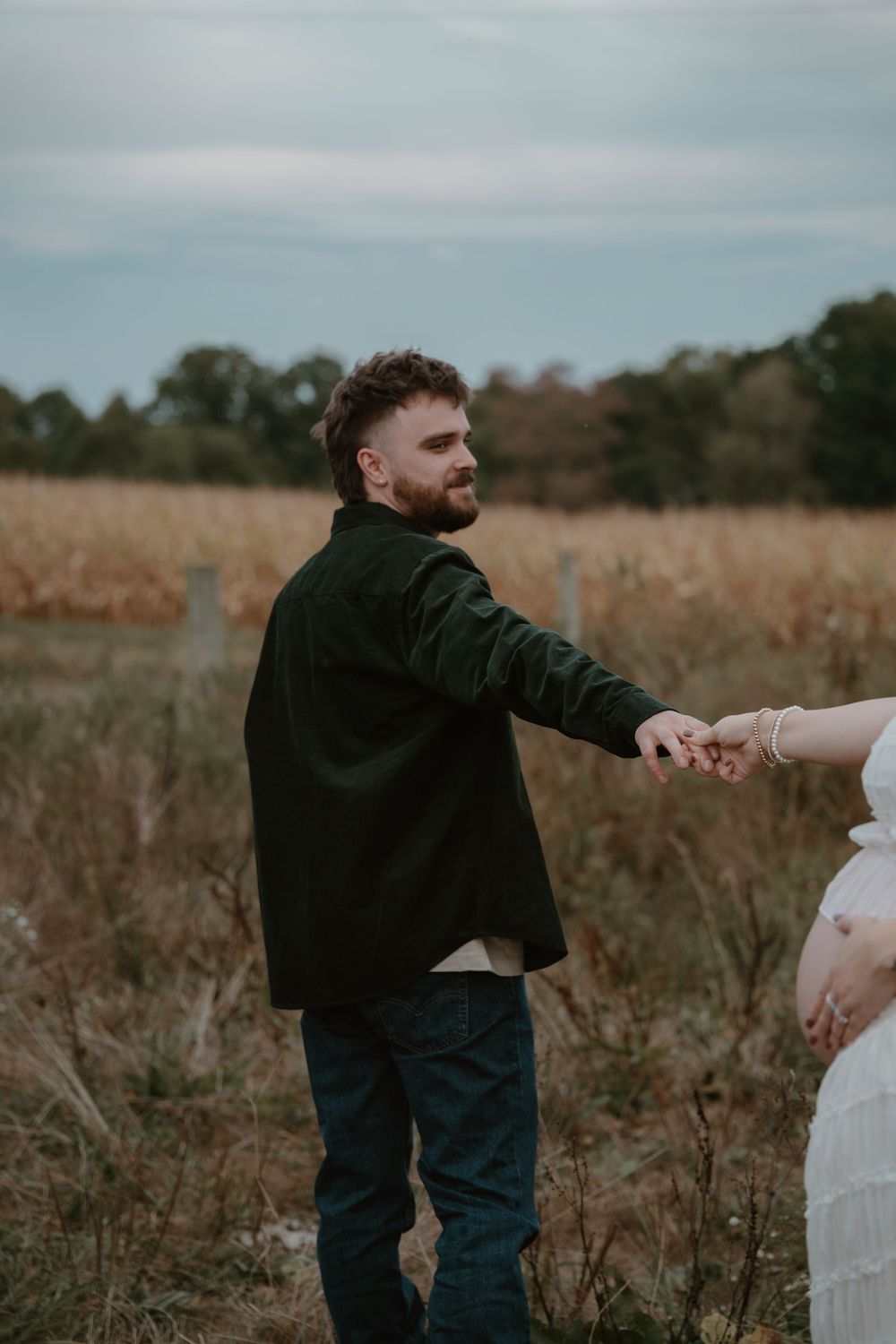 Man in dark shirt holds pregnant woman's hand in golden field during outdoor maternity photoshoot.