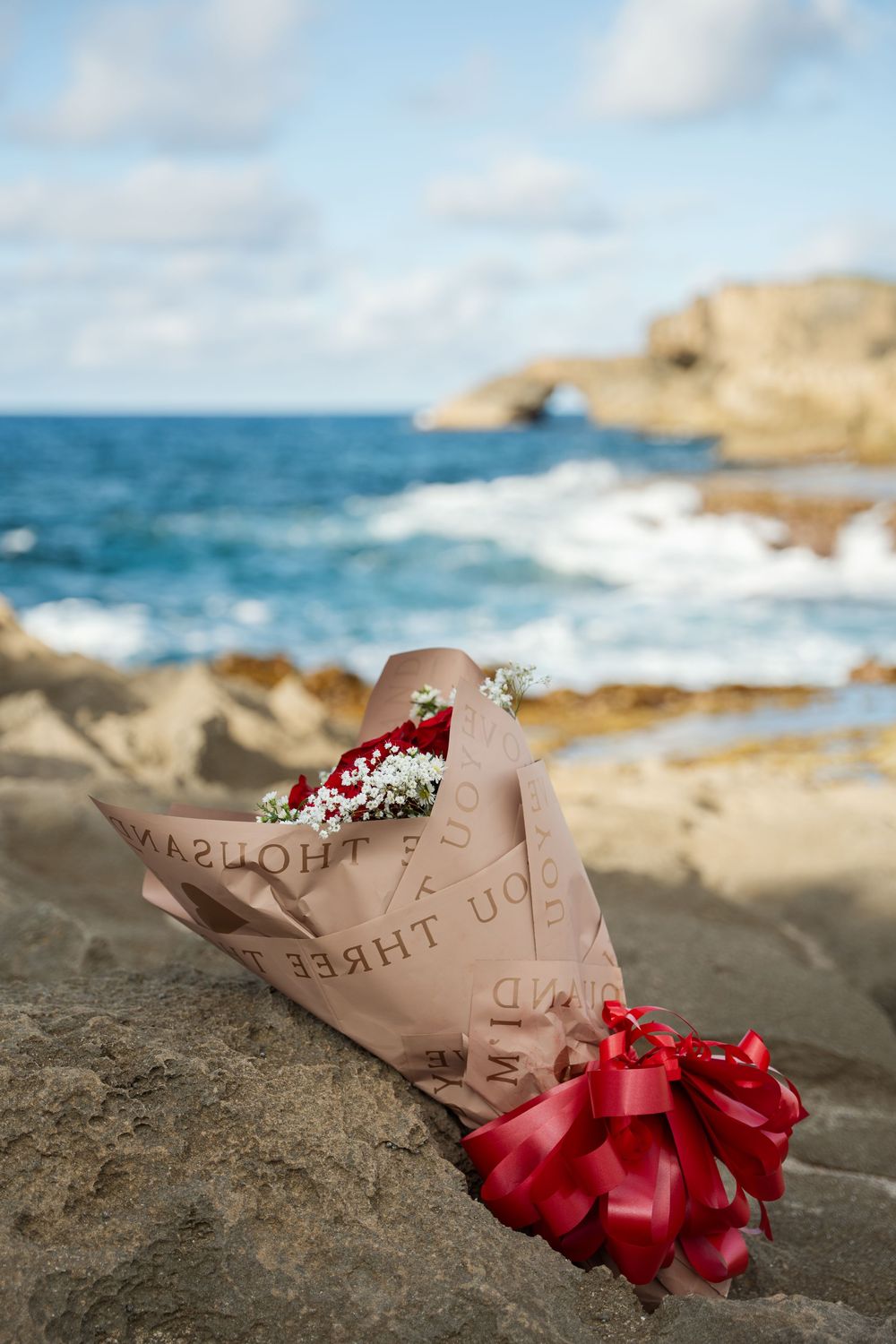 A wrapped bouquet with red flowers sits on a rocky coastal shore with blue ocean waves and cliffs in the background.