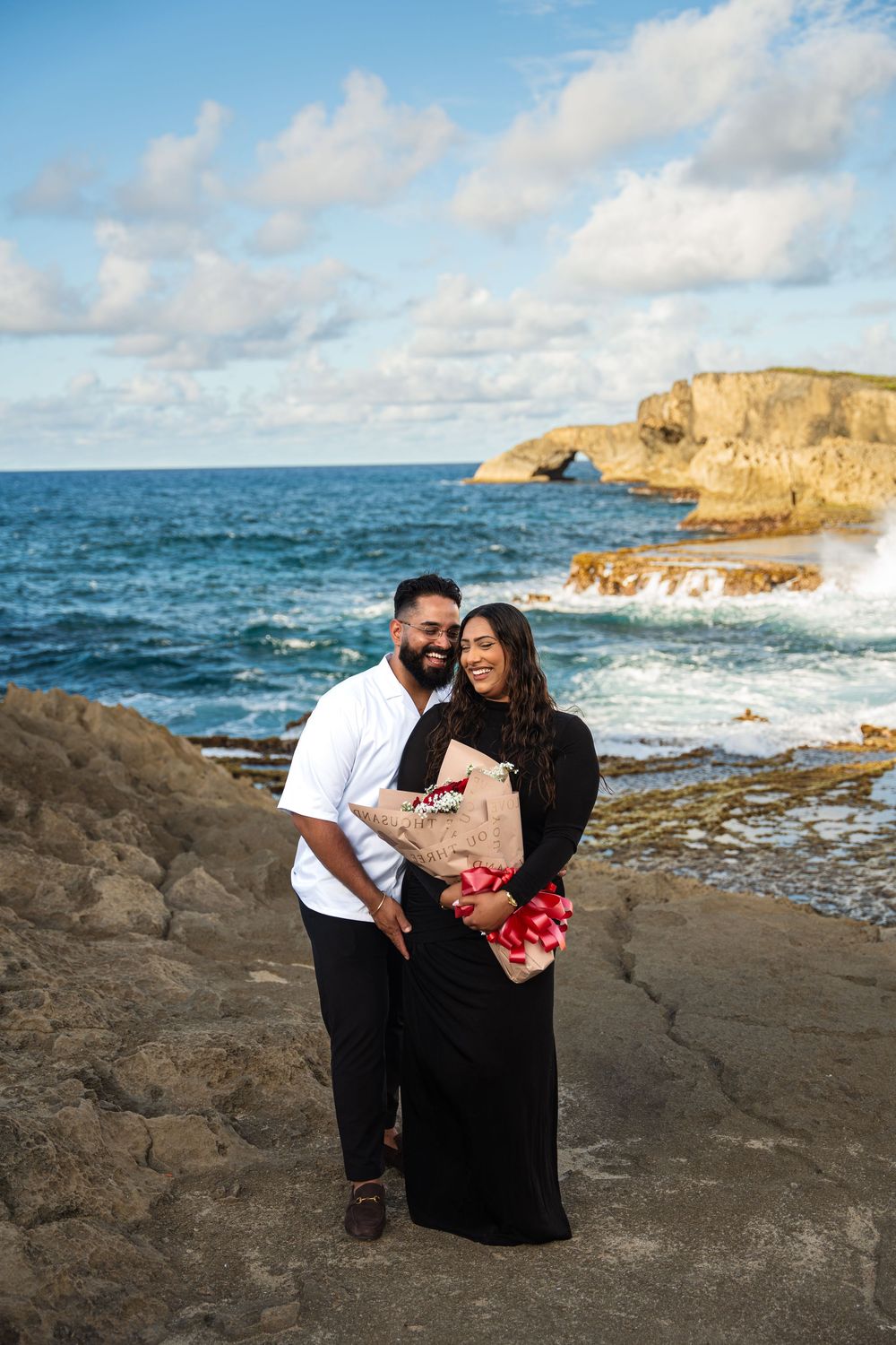 Couple sharing romantic moments on rocky coastal cliffs with ocean waves and blue sky in the background.