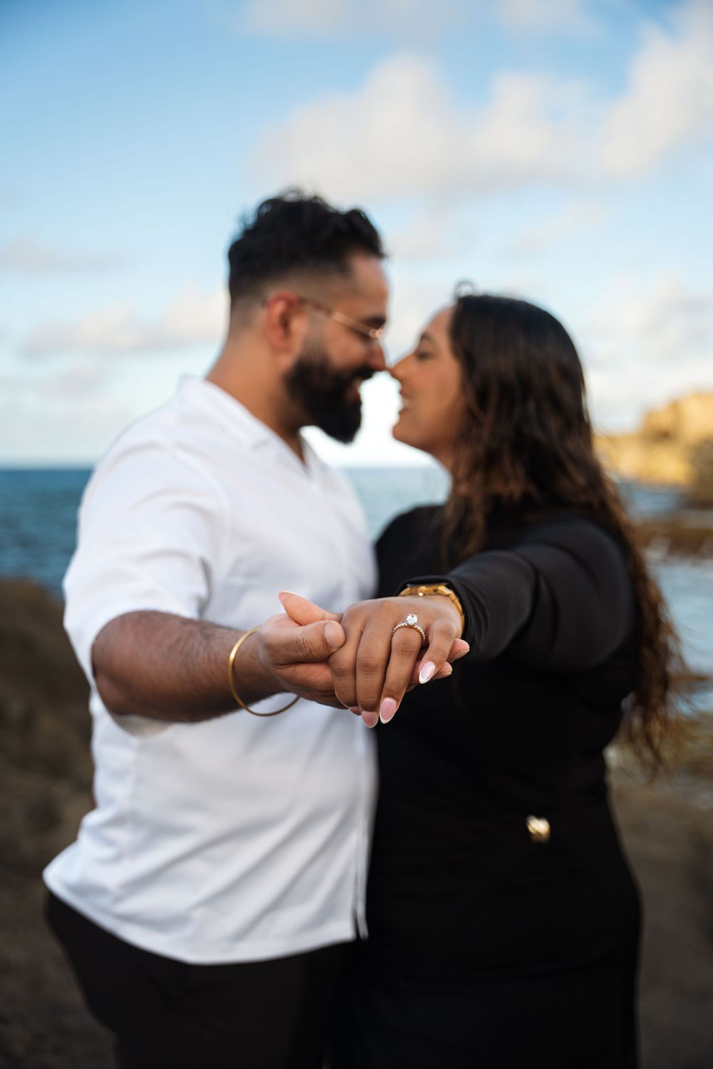 Couple showing engagement ring against dramatic coastal backdrop with crashing waves and cliffs.