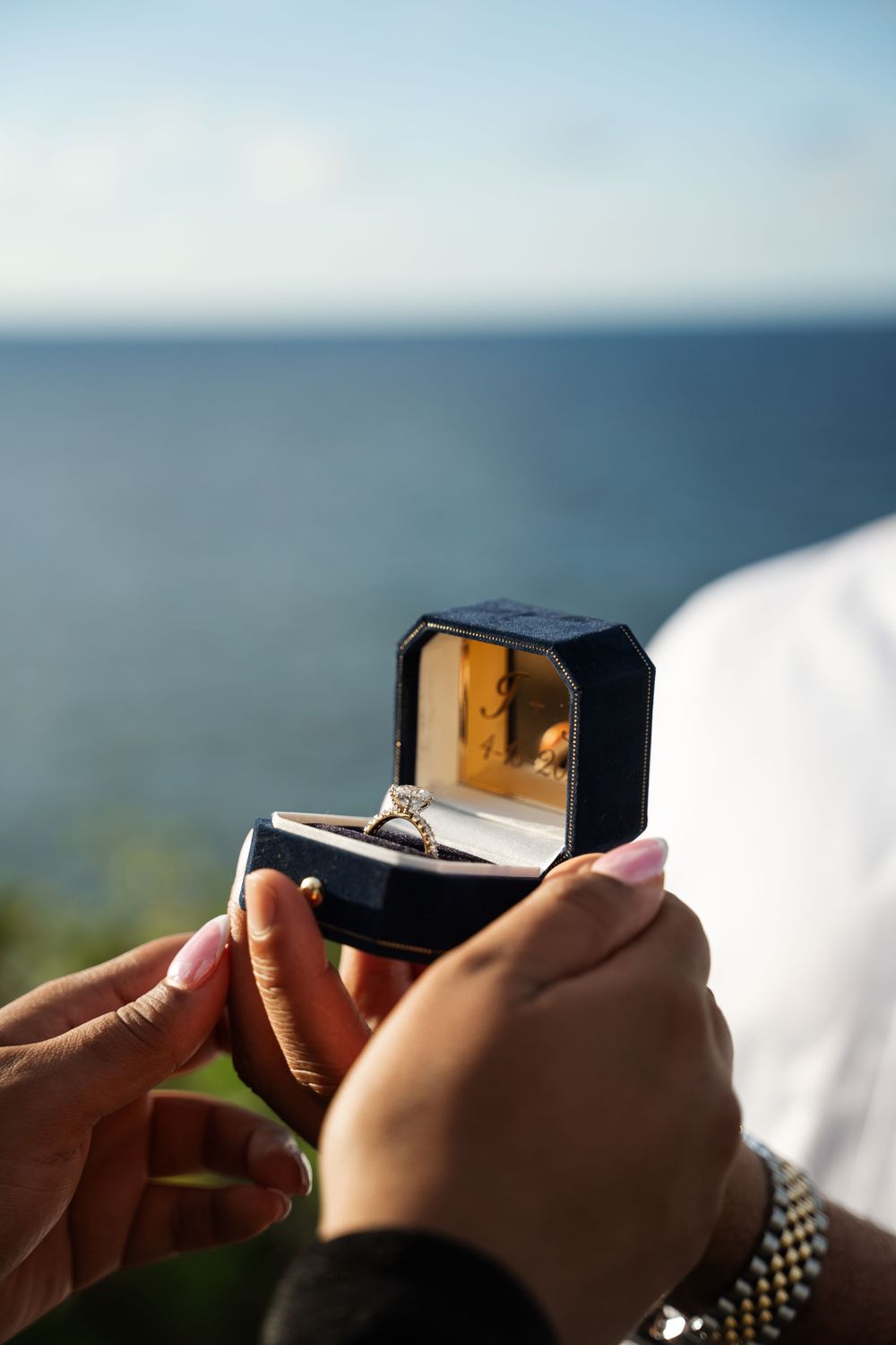 Hands holding a ring box with a diamond engagement ring against a blurred ocean background.