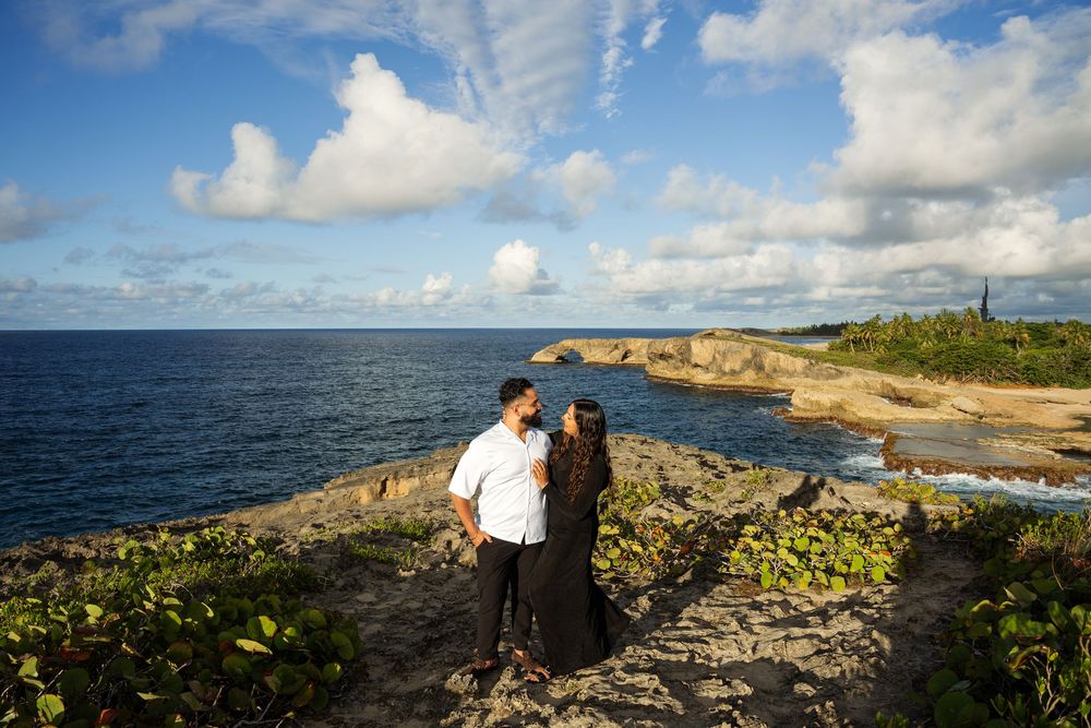 Couple stands on rocky coastal cliffs overlooking blue ocean waters and dramatic cloudy sky at sunset.