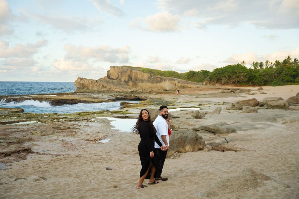 Two people stand together on rocky coastline with cliffs and ocean waves at sunset in Hawaii.