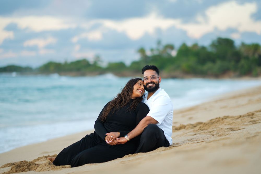 A couple in black outfits lies romantically on a sandy beach during a photo session at sunset.