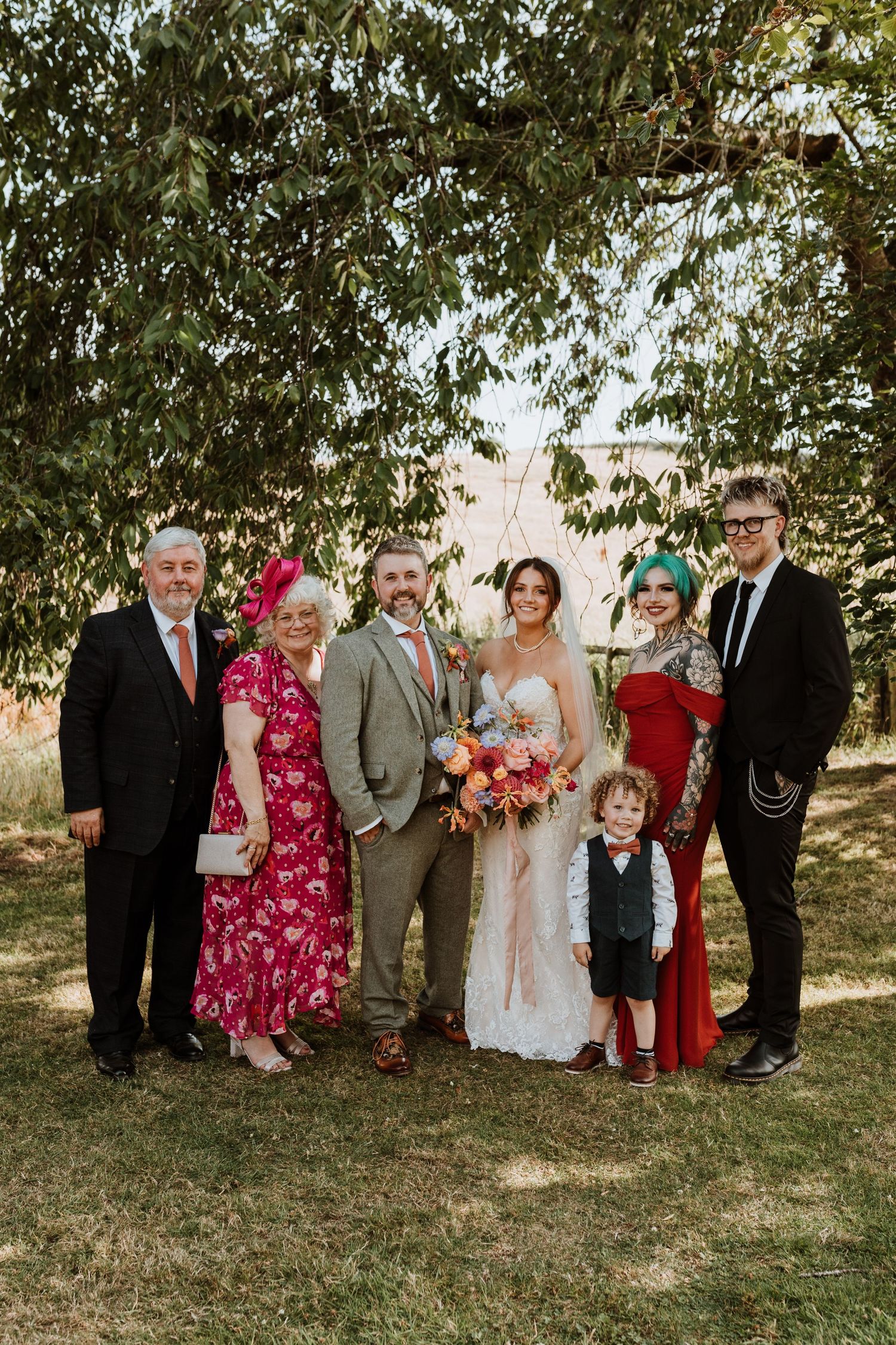 Family group poses outdoors under green trees at wedding with bride in white dress and colorful floral bouquet at Lyde Court