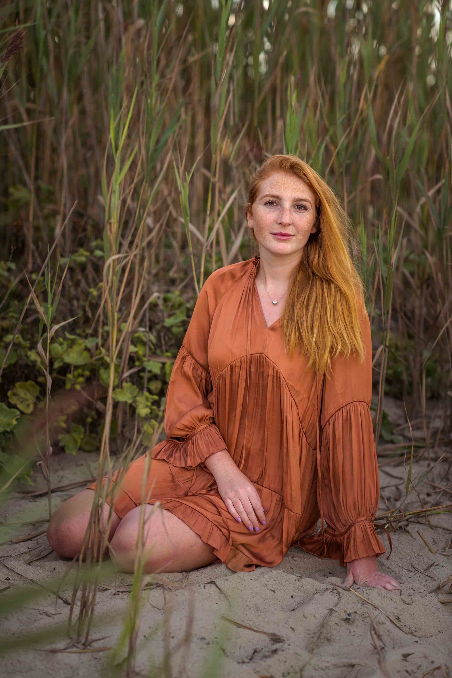 Young redhead woman in a rust-colored dress sitting barefoot on sandy ground among tall green reeds.