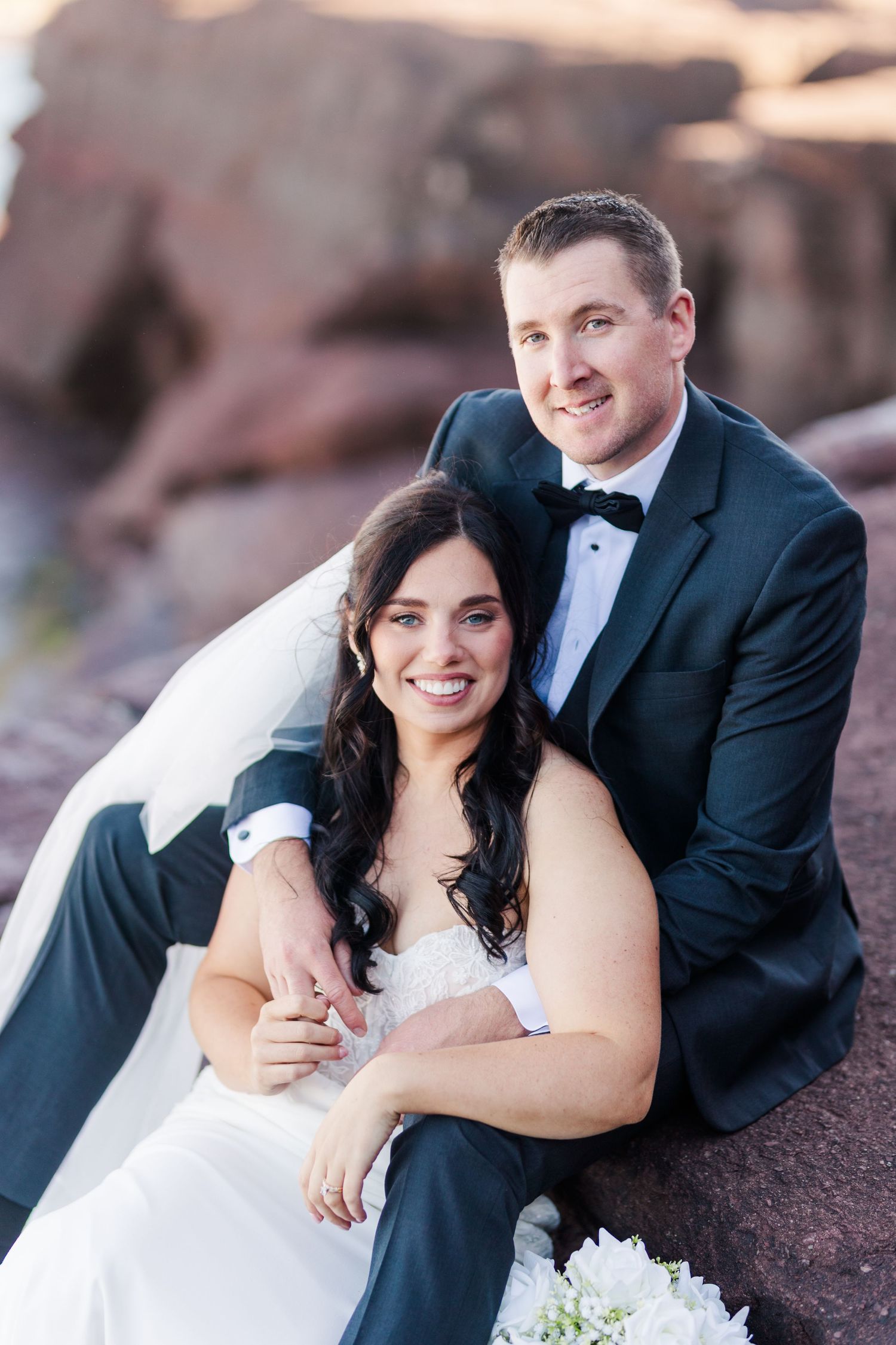 A bride in white dress and groom in dark suit pose together on rustic rocks during outdoor wedding photoshoot.