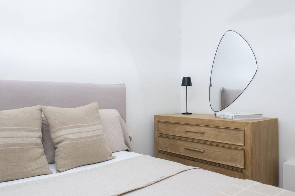 Modern minimalist bedroom interior with beige bedding, wooden dresser and oval mirror on white walls.