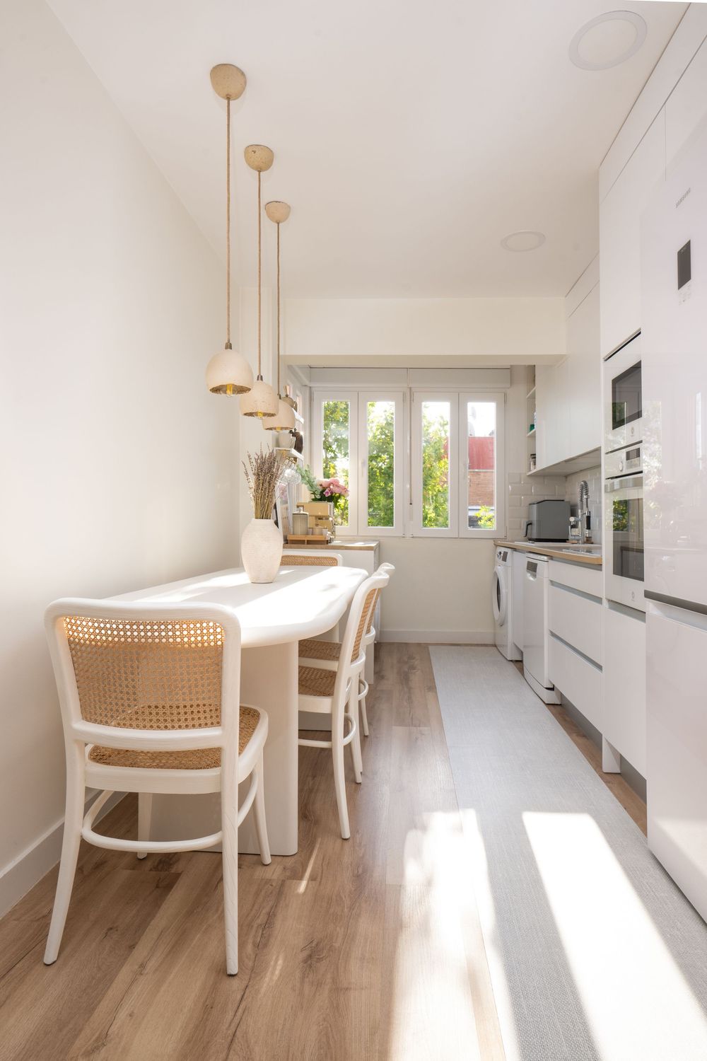 Modern white kitchen with wood floors and cane dining chairs features pendant lights and abundant natural lighting.