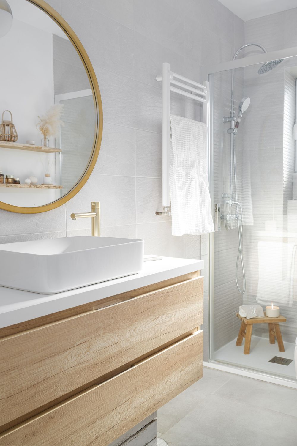 Modern white bathroom interior with round gold mirror, wood vanity, and glass shower enclosure.