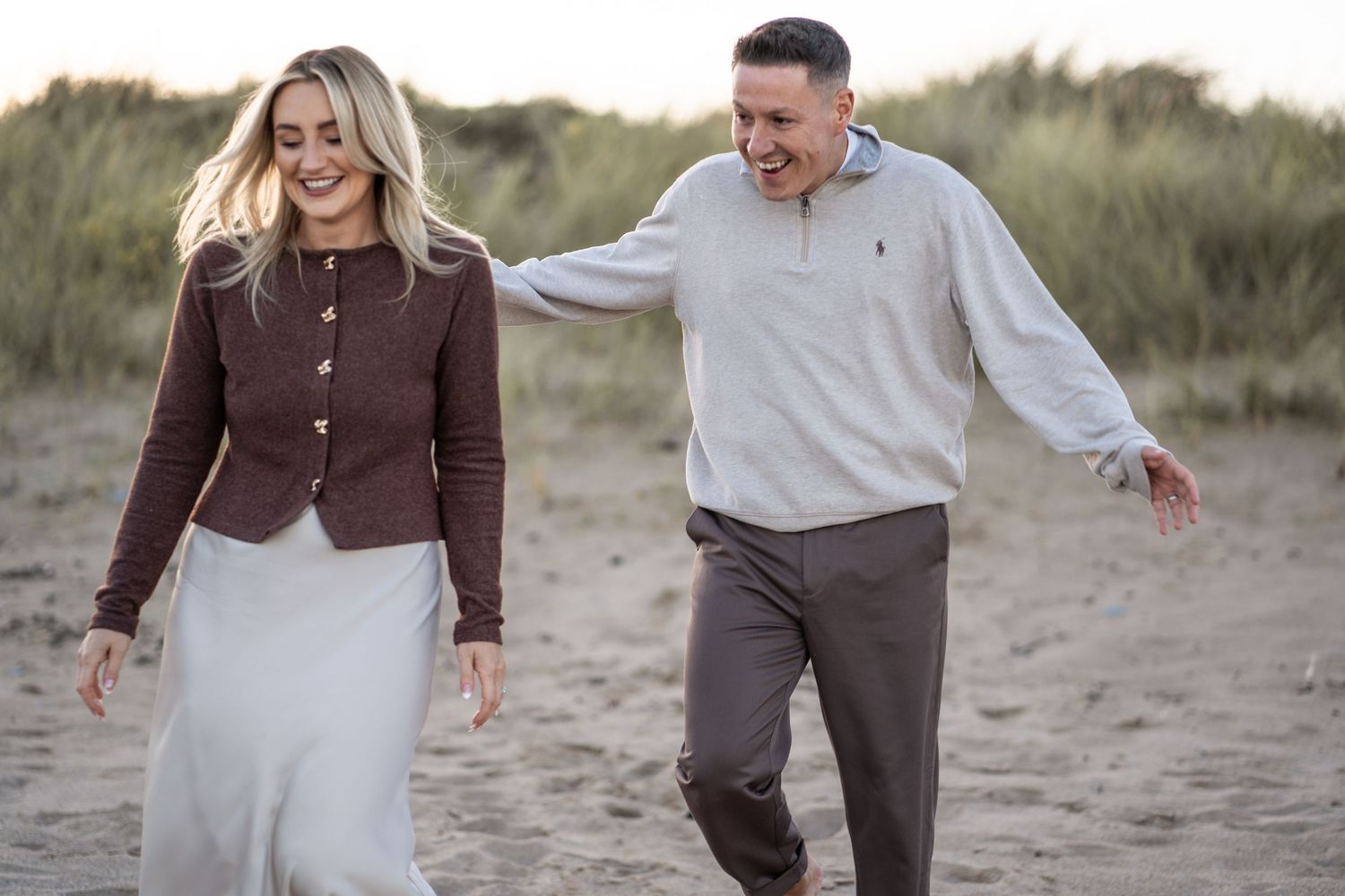 A couple laughs and plays together on a sandy beach during a romantic sunset photoshoot.