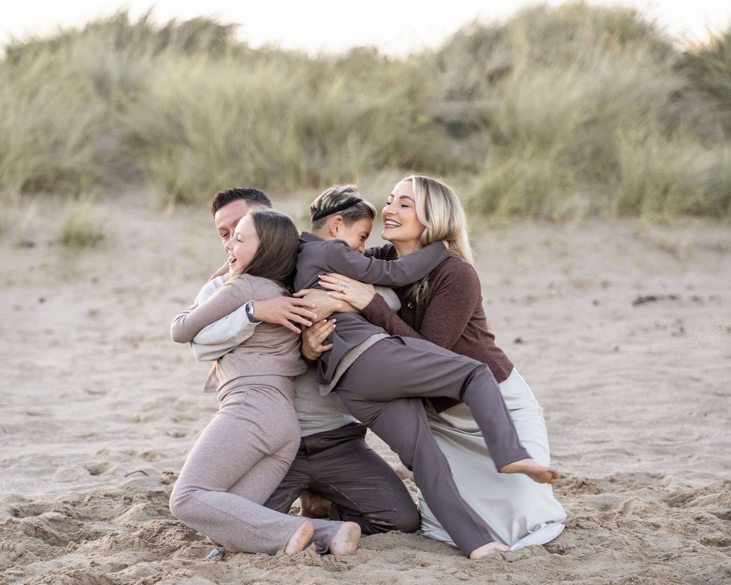 A group embraces while sitting together on a sandy beach with dunes in the background.