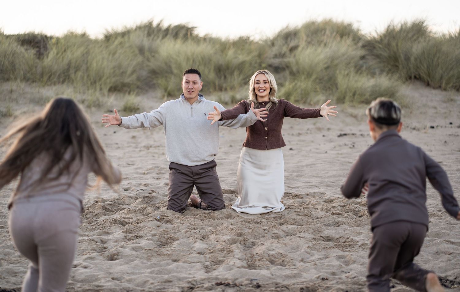 A couple poses playfully on a sandy beach with tall grass dunes in the background while others take their photo.