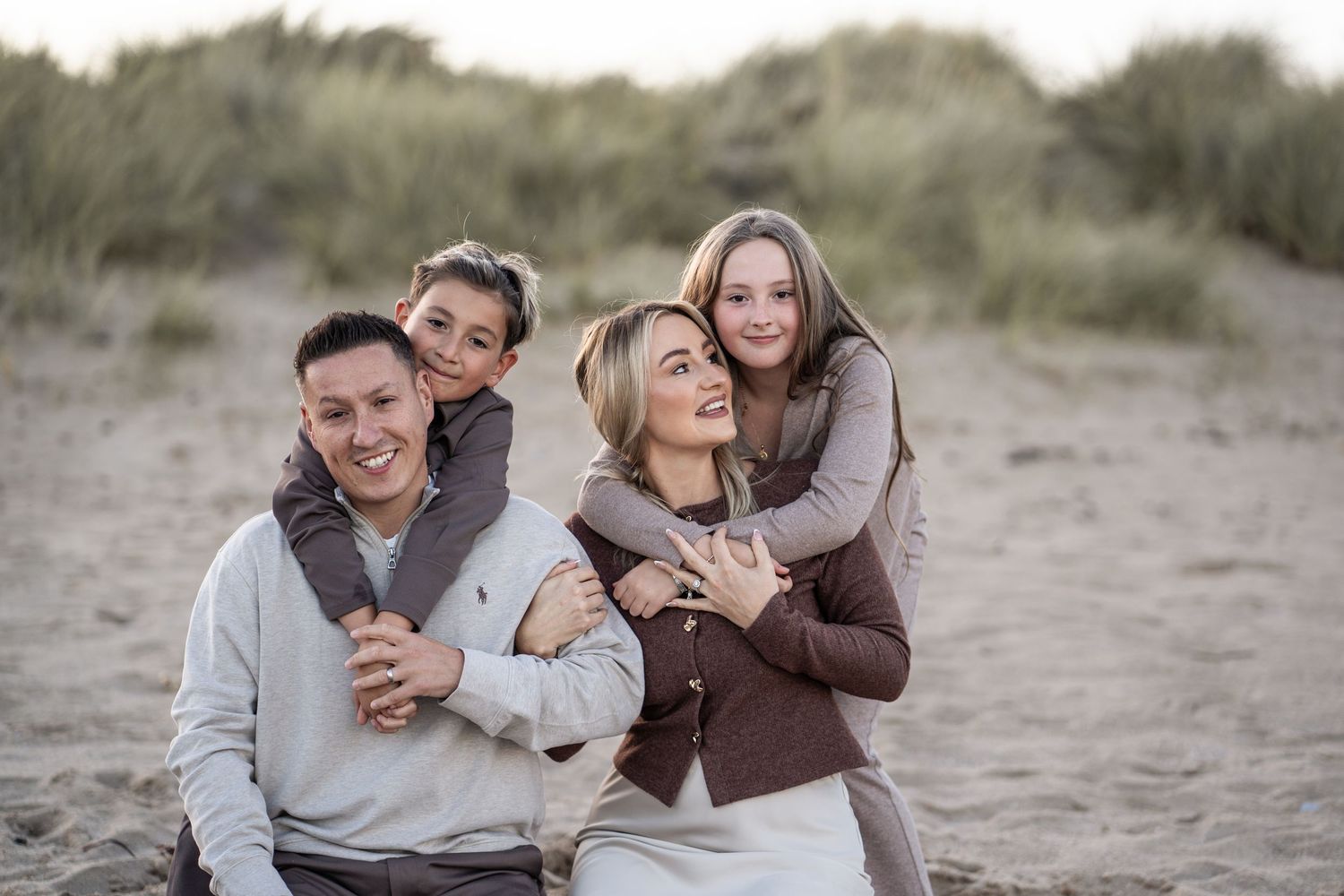 A couple embraces on a sandy beach during sunset wearing neutral toned clothing.