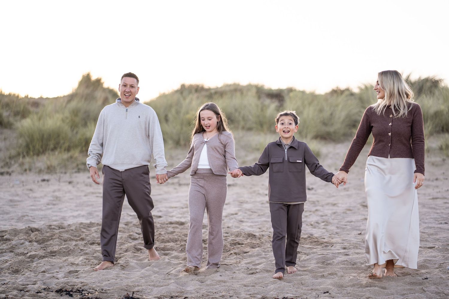 Group walking hand in hand along sandy beach path at sunset with dunes in background.