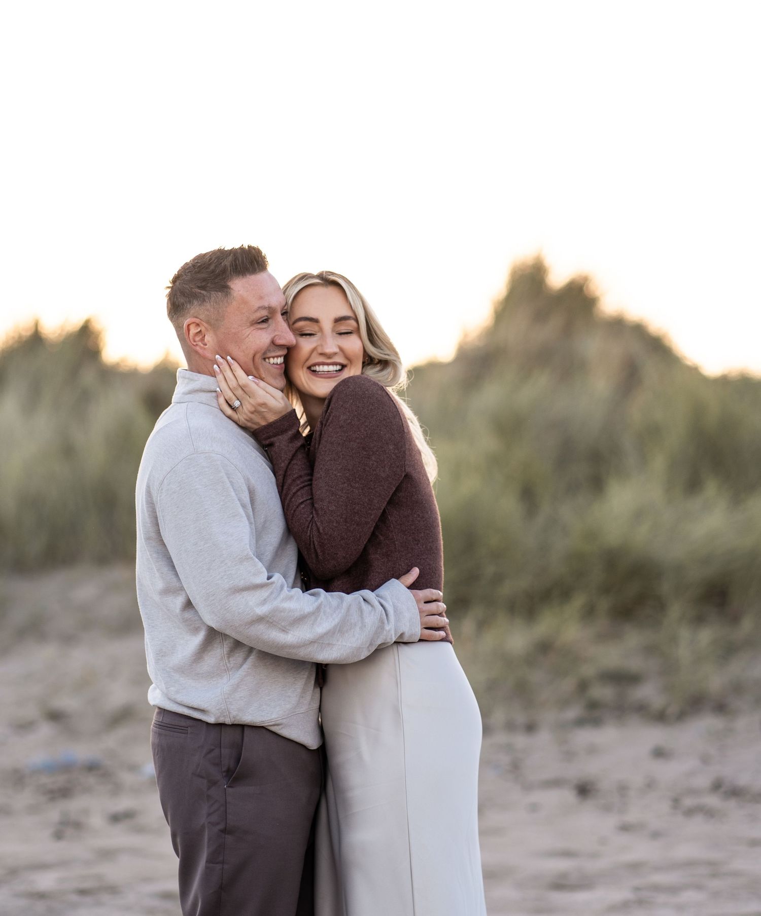 Couple sharing intimate moment on beach with sand dunes and golden sunset light in background.
