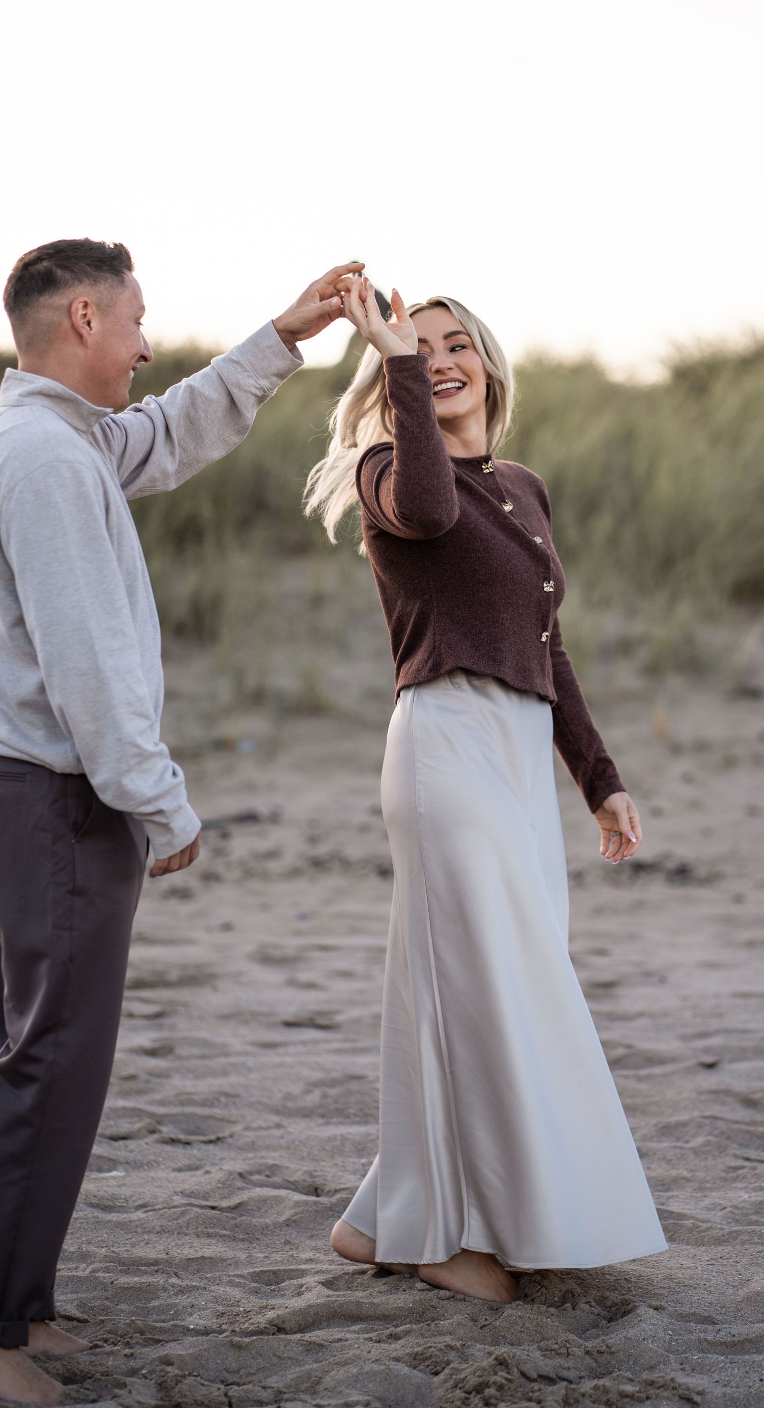 A couple dances joyfully together on a sandy beach at sunset in casual attire.