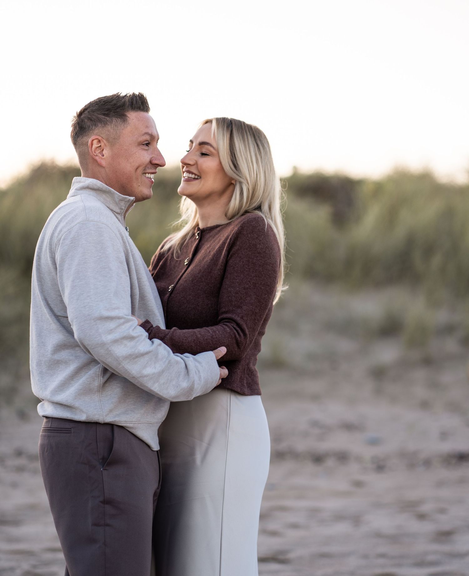 Couple embracing on a beach at sunset wearing neutral colored sweaters.