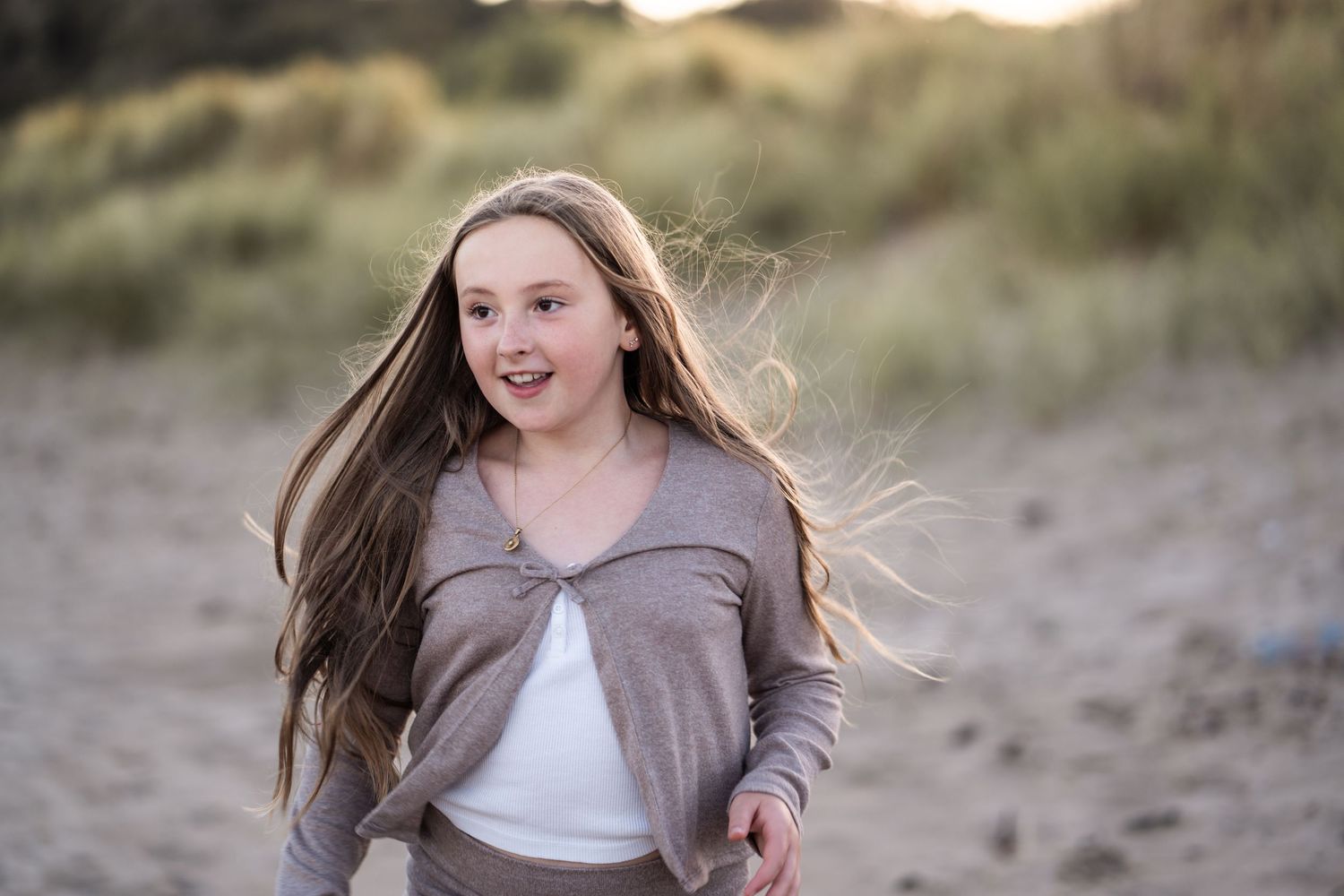 A person with long brown hair wearing a gray sweater and white top smiles while walking on a sandy beach at sunset.