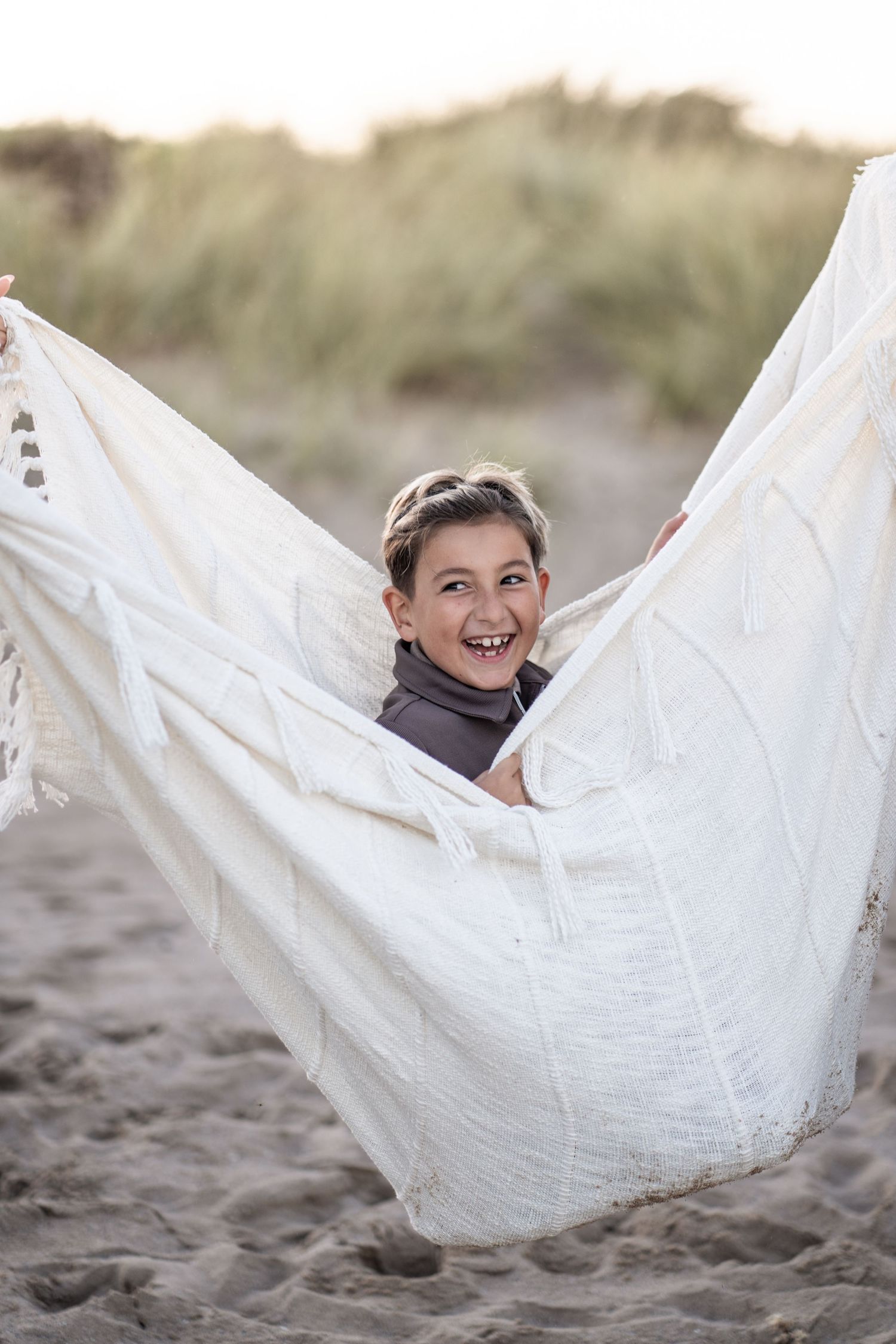 Child wrapped in white fabric smiling joyfully while playing at the beach on a sunny day.