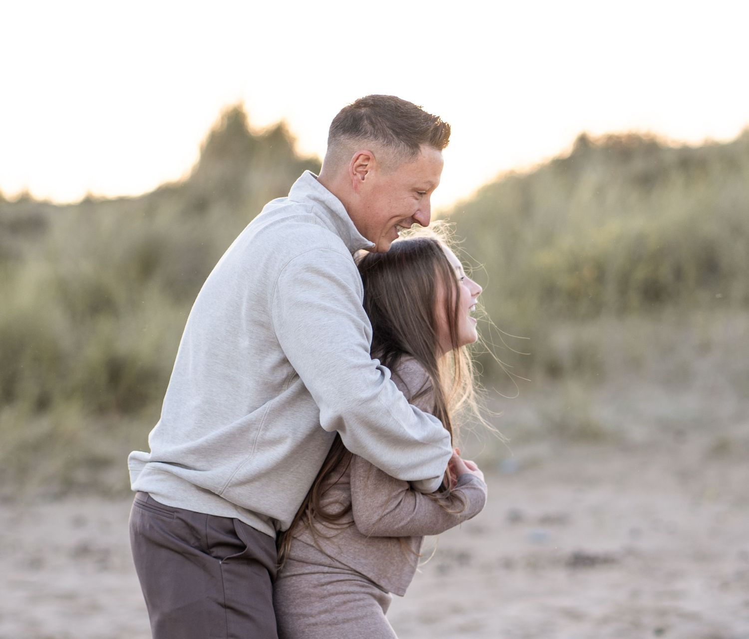 A couple shares an intimate moment while sitting together on a beach with dunes in the background.