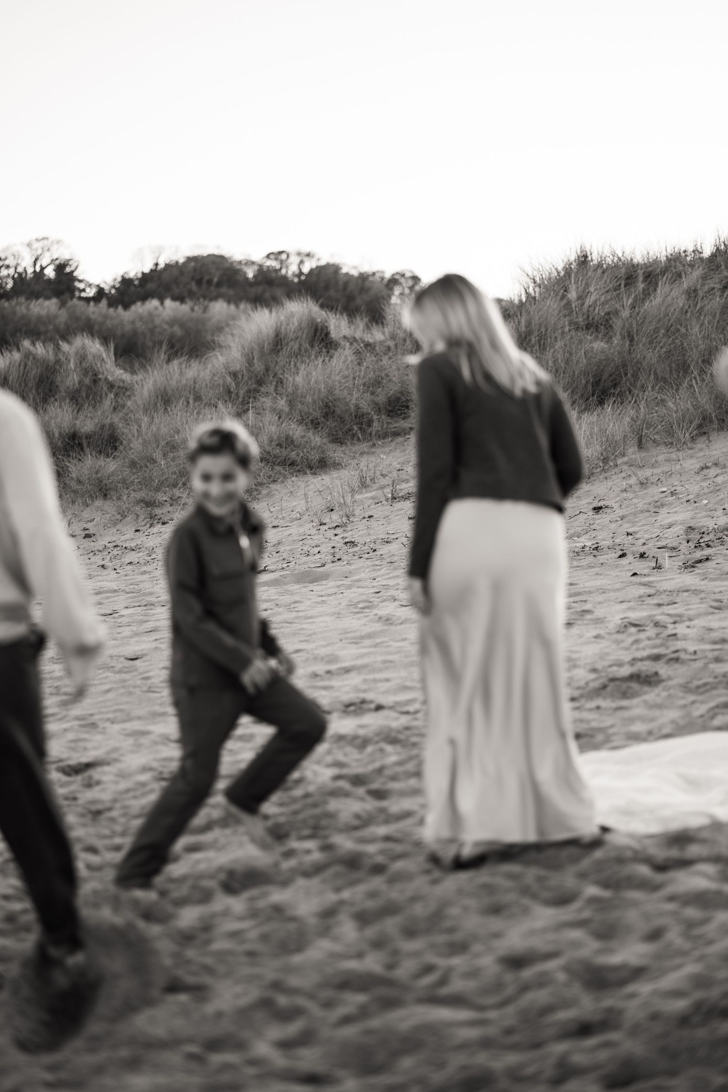A black and white beach scene showing people walking on sandy dunes with grass in the background.