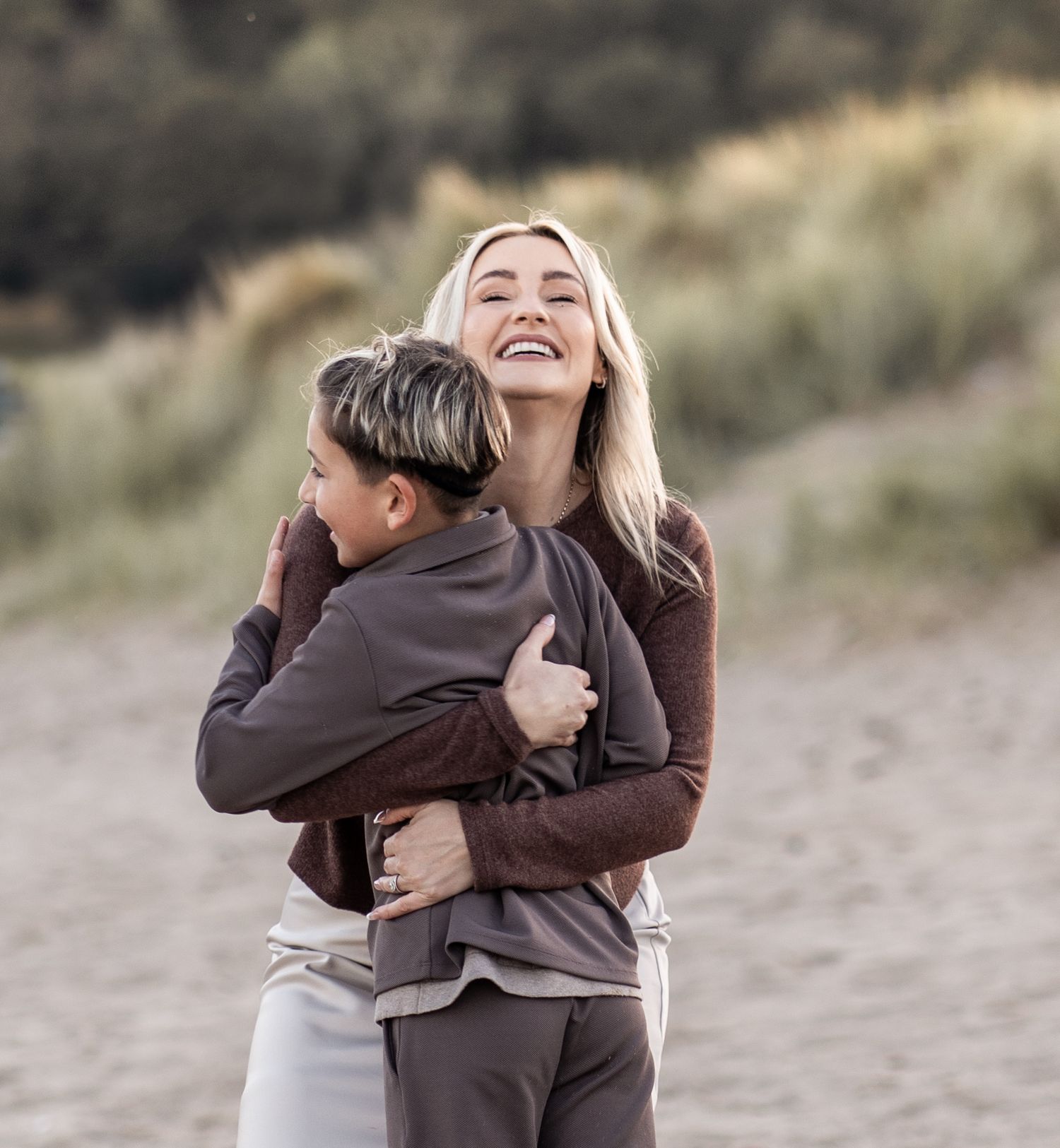 People embrace and laugh together on a sandy beach path surrounded by dune grass.