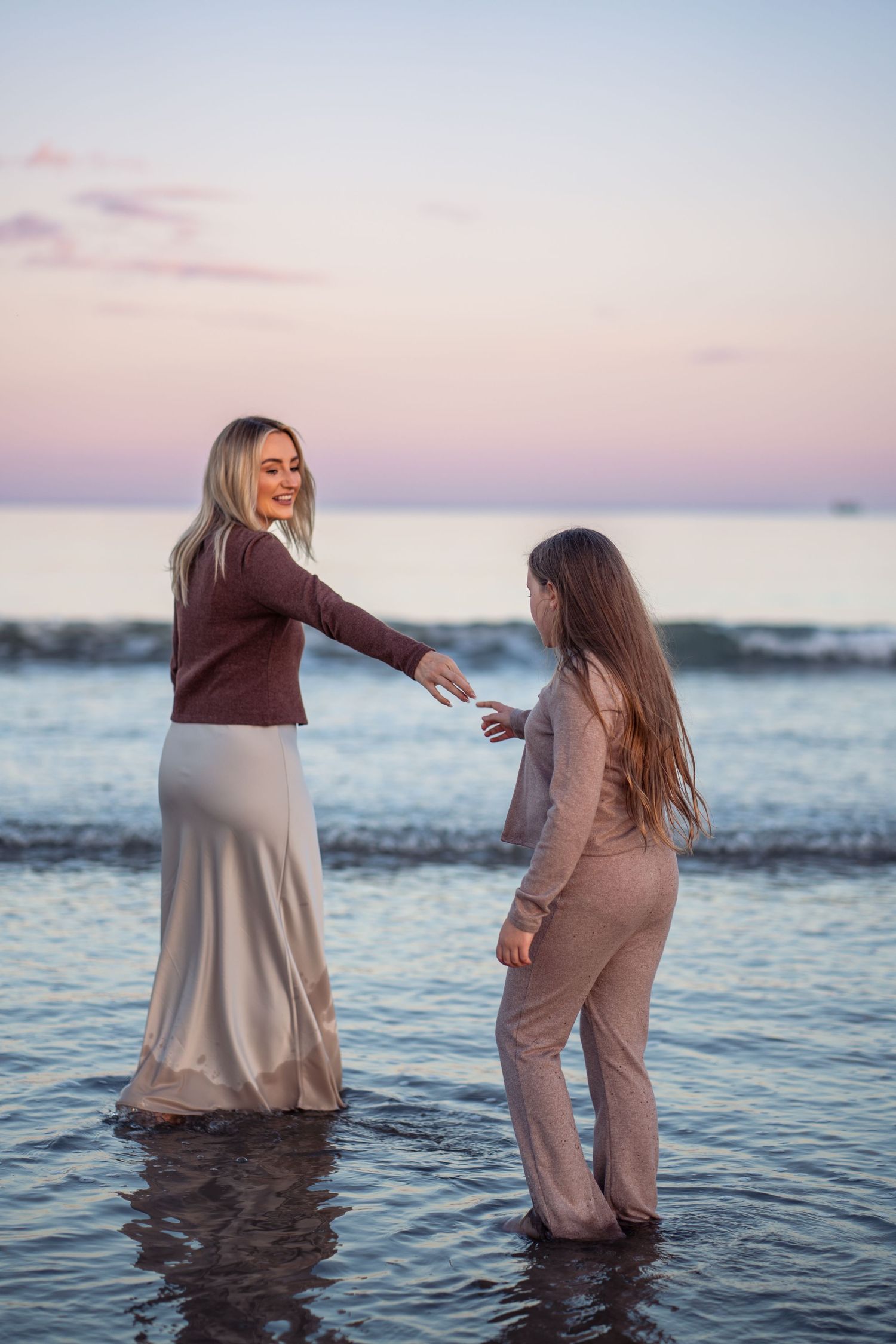 Two people stand together in shallow ocean water at sunset creating a romantic silhouette against the pink sky.