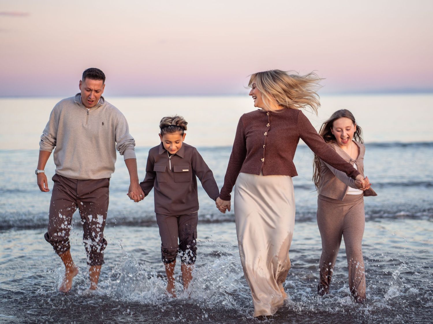 Family members walk together through shallow ocean waves at sunset creating joyful splashes.