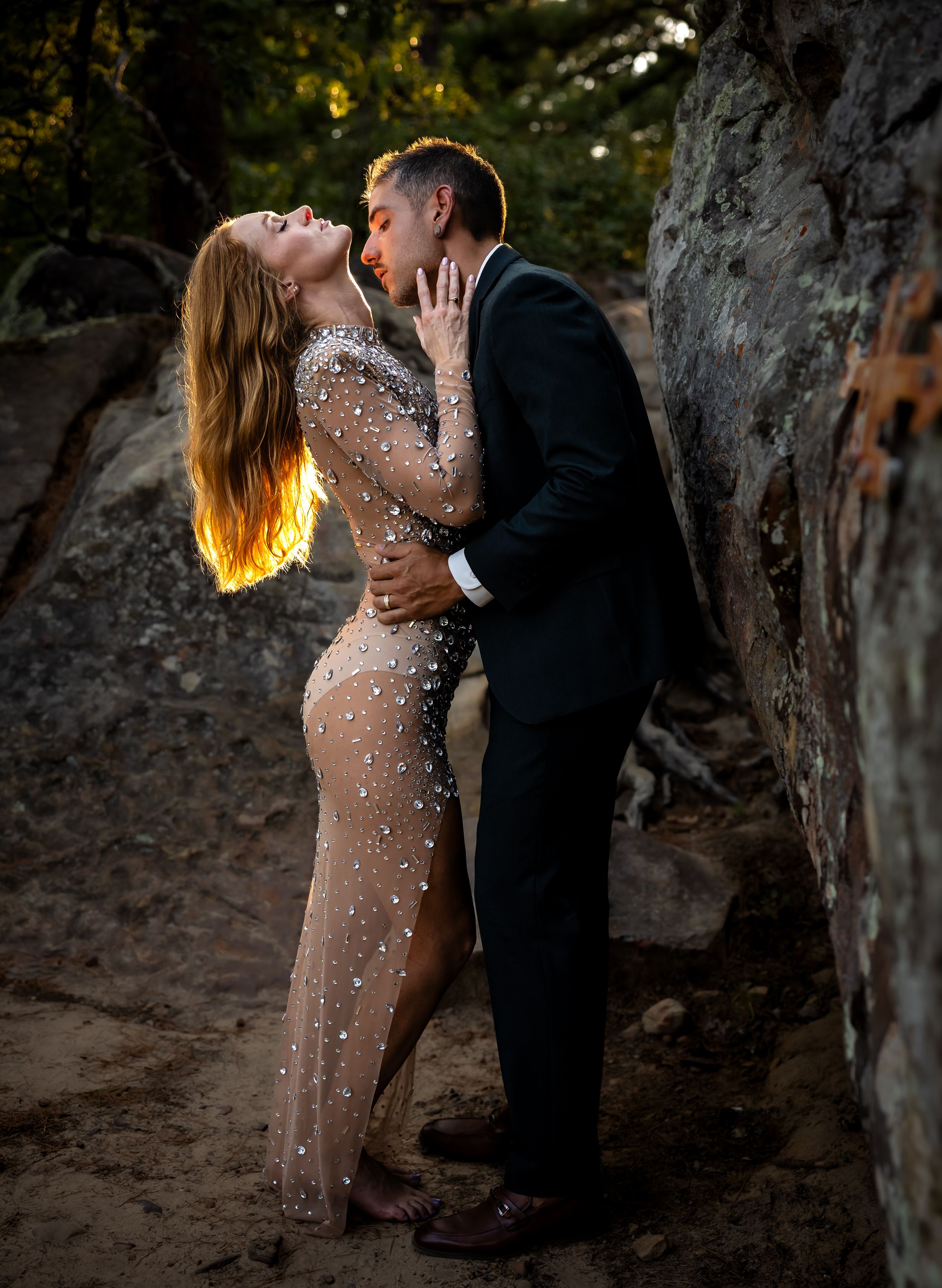 Romantic couple embraces in a sheer sparkly dress and suit among trees at dusk.