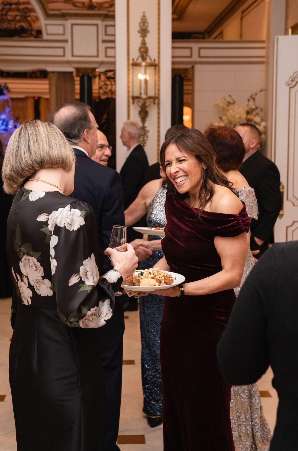 Elegantly dressed guests socializing at a formal gala event, woman in burgundy velvet off-shoulder gown holding appetizer plate.