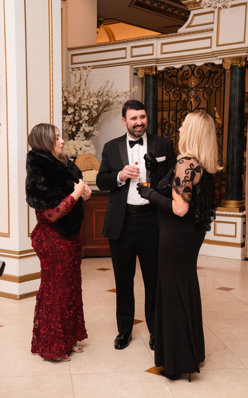 Elegant guests in formal attire chat and mingle at an upscale event in a luxurious marble-columned venue.