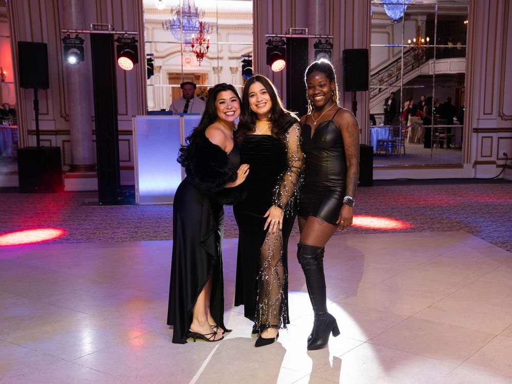 Dancers in black outfits practicing steps together on a polished dance floor in a ballroom with pink lighting.