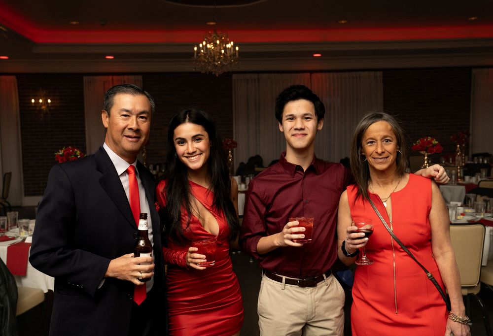 Group of people in red attire holding drinks at a social gathering with warm lighting and chandeliers.